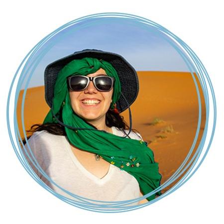 A student wearing a green scarf, sun hat, and sunglasses smiles while standing in a Moroccan desert