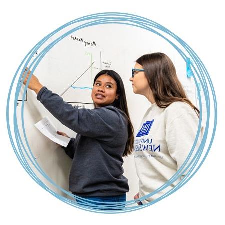 A student points to and discusses an economics graph on a white board with another student