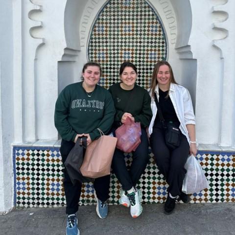 Three female students pose in front of Moroccan architecture. Photo is set against a blue background.